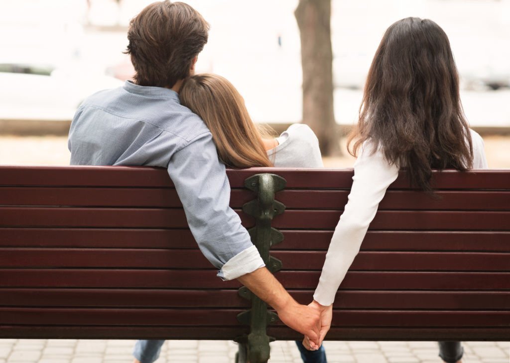 Love Triangle. Cheating Boyfriend Hugging Girlfriend Holding Hands With Her Girl Friend Sitting On Bench Together In Park Outdoor. Back-View